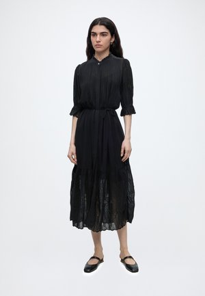 Femme aux longs cheveux foncés portant une robe midi noire à manches trois-quarts, une ceinture à la taille et des ballerines Mary Jane noires, immobile.