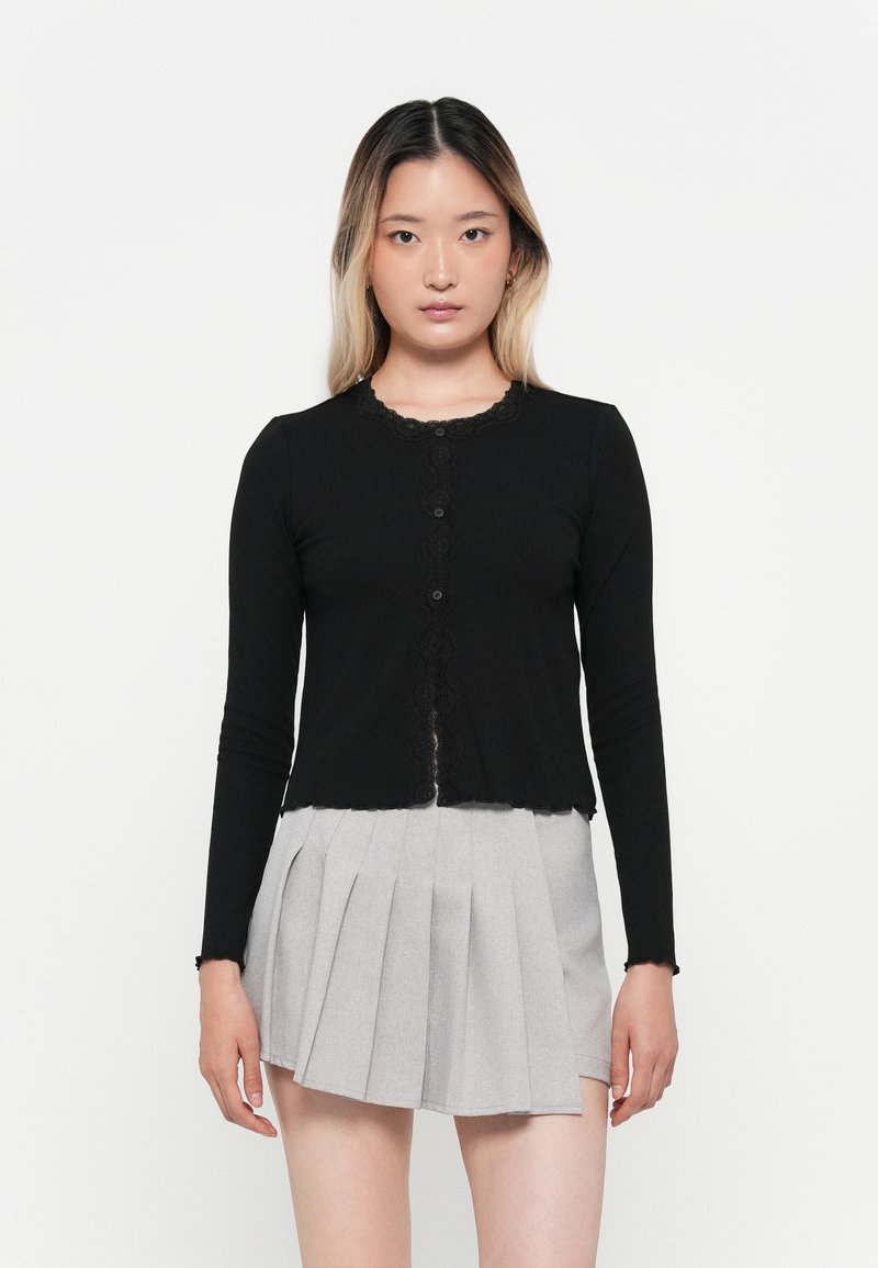 Svart langermet cardigan med blonder og frontknapper, kombinert med et lys grått plissert skjørt. Glatt tekstur og tettsittende design.