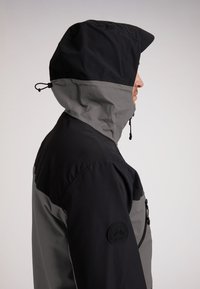 Giacca con cappuccio in una combinazione di colori nero e grigio. Presenta un tessuto testurizzato, un cappuccio regolabile e una tasca laterale con zip dotata di un'emblema.