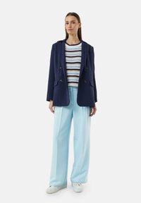 Granatowa marynarka dwurzędowa, paskowany sweter z dzianiny, jasnoniebieskie szerokie spodnie, białe sneakersy. Gładkie faktury i dopasowany krój.