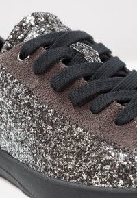 Gros plan d'une basket avec des lacets noirs, des détails en daim marron et des paillettes argentées couvrant le côté et la languette.