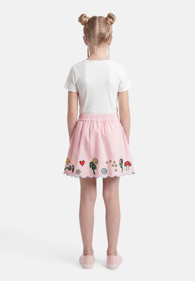 Κορίτσι με ξανθά μαλλιά σε δύο κότσους φοράει λευκό τοπ με κοντά μανίκια και ροζ φούστα με κεντημένα λουλούδια, αστέρια, καρδιές και μανιτάρια, όρθια.