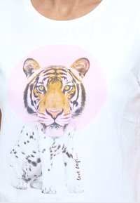 Visage de tigre combiné avec le corps d’un chien tacheté, assis au centre d’un t-shirt blanc avec un cercle rose pâle derrière la tête du tigre.