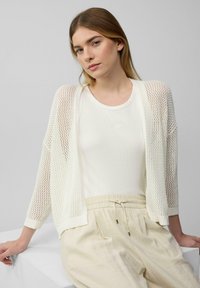 Witte opengehaakte cardigan over een crèmekleurige top, gecombineerd met lichtbeige broek. Losse pasvorm, gestructureerd breiwerk, met een casual, gelaagde uitstraling.
