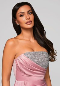 Kvinna med långt mörkt hår, iförd en ärmlös rosa klänning med en glittrande silverpärlade bodice, mot en enfärgad ljusgrå bakgrund.