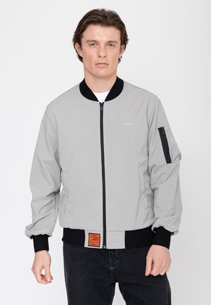 Młody mężczyzna w jasnoszarej kurtce bombowej z czarnymi mankietami, kołnierzem i zamkiem błyskawicznym, stojący na tle jednolitego białego tła.