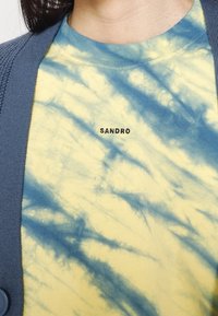Gul och blå tie-dye bomulls t-shirt med rund halsringning och broderade ordet "SANDRO" i svart, lager på lager under en grå cardigan.