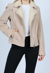 Frau in einer beigen Wildlederjacke mit Lammfellkragen, weißem Hemd und schwarzen Hosen, mit einer Hand in der Jackentasche.