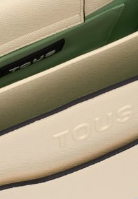 Jasno beżowa torebka z teksturowanej skóry, z zielonym wykończeniem wnętrza. Posiada wytłoczone logo "TOUS" oraz kontrastowe czarne wykończenia na brzegach.