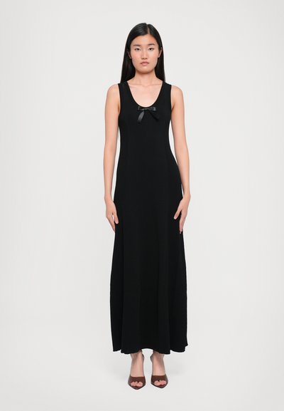 Zwarte, mouwloze maxi-jurk met geribbelde textuur en een strikaccent op de borst. Model draagt bruine open teen hakken.