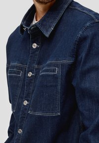 Donkerblauw denim overhemd met knoopsluiting, twee borstzakken en zichtbare stikdetails. De stof lijkt textuur en duurzaam te zijn.