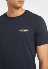 Námořnické bavlněné tričko s kulatým výstřihem, krátkými rukávy a červenobílým logem "GUESS JEANS" nad levou částí hrudníku.