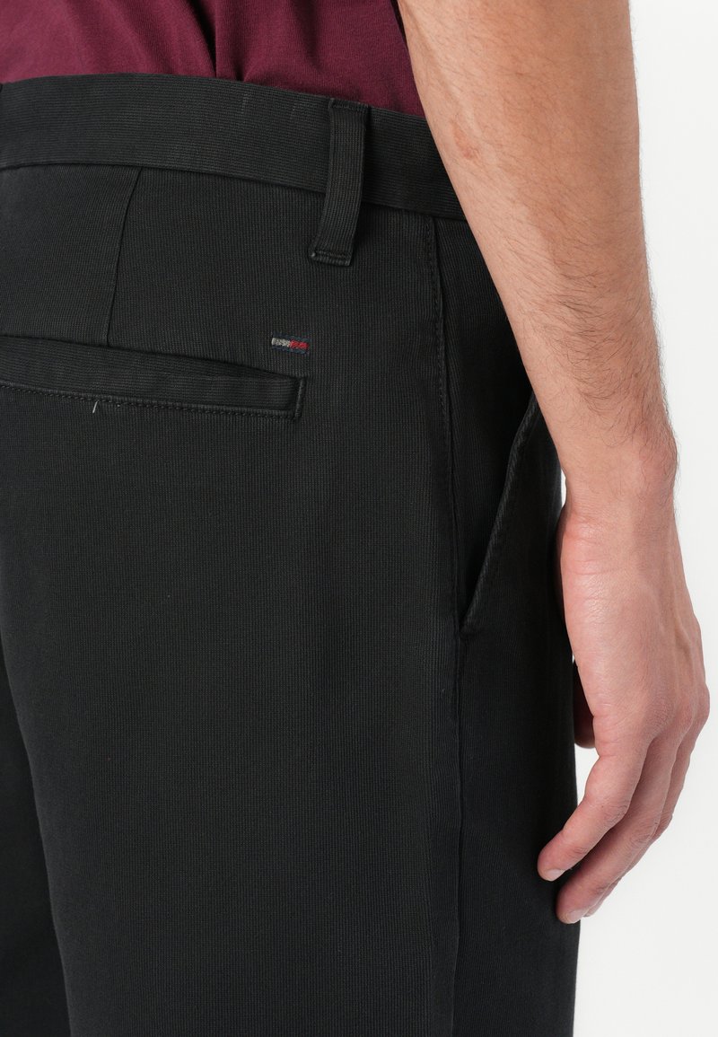 Schwarze Baumwollhosen mit ribbeliger Textur, ausgefeilten Nähten, seitlichen Taschen und einem kleinen Logotaschenetikett in der Nähe der Gesäßtasche.