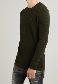 Maglione texture verde scuro con scollo rotondo, dotato di una piccola toppa del logo nero. Indossato con jeans grigi strappati.
