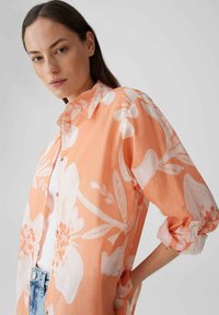 Camicia color pesca con motivi floreali bianchi, con bottoni sul davanti e maniche arrotolate. Realizzata in tessuto leggero con una vestibilità comoda.