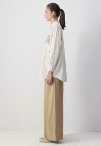 White oversized shirt with button closure and floral embroidery on the chest, paired with beige wide-leg pants and light footwear.