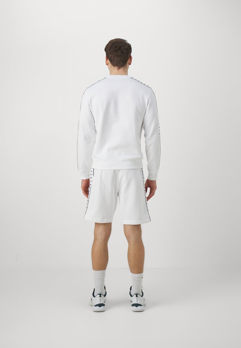 Completo in felpa bianca con pantaloni corti, caratterizzato da polsini a coste. Presenta strisce laterali nere. Il tessuto sembra morbido e strutturato. Scarpe da ginnastica completano l'outfit.