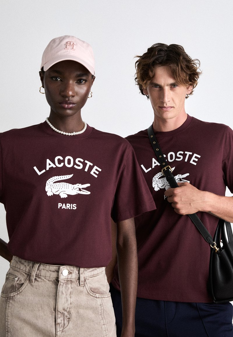 Camisetas color burdeos con un logotipo blanco de Lacoste y un gráfico de cocodrilo. Un modelo lleva un collar de perlas, el otro sostiene un bolso negro con una correa.