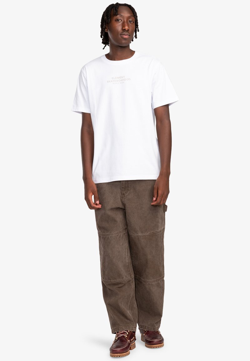 T-shirt blanche à manches courtes avec impression de logo, associée à un pantalon en velours côtelé brun ample et des chaussures brunes avec lacets.