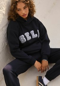 Marineblauer Kapuzenpullover mit dem Wort "SELF" in hellgrau über der Brust, kombiniert mit passenden Hosen. Weicher Stoff und lockere Passform.