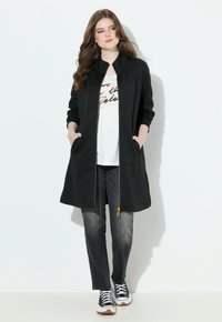 Mujer vestida con una chaqueta larga negra con cremallera, camiseta blanca con estampado, jeans negros desgastados y sneakers en blanco y negro, de pie contra una pared blanca.