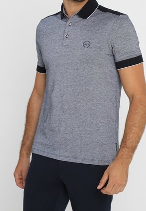 Mies, joka on pukeutunut harmaaseen lyhythihaiseen poolopaitaan, jossa on musta kaulus ja hihansuut, sekä tummiin housuihin, seisoo yksiväristä taustaa vasten.