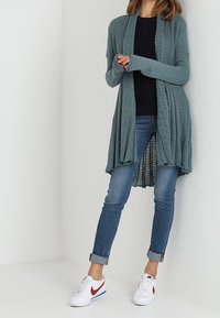 Kvinna i en teal öppenstickad kofta, svart topp, upprullade blå jeans och vita Nike-sneakers med röd swoosh-logotyp, stående mot en vit vägg.