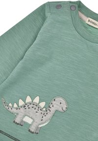 Maglietta verde a maniche lunghe con ricamo di un dinosauro grigio, dotato di spine e macchie, realizzata in tessuto testurizzato e con bottoni a pressione sulla spalla.