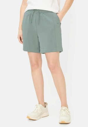 Osoba ubrana w niebiesko-zielone szorty z troczkami, biały top i białe tenisówki, stoi z jedną ręką w kieszeni na białym tle.