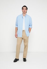 Ανοιχτό μπλε και λευκό ριγωτό πουκάμισο με κουμπιά φορεμένο πάνω από λευκό μπλουζάκι, σε συνδυασμό με μπεζ παντελόνια και μαύρα παπούτσια. Απλός, καθημερινός σχεδιασμός.