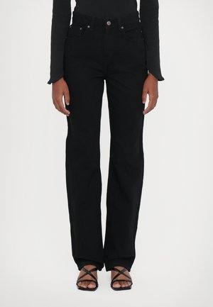Black high-waisted jeans with a straight leg cut. Features five pockets, a button closure, and subtle stitching detailing. Paired with black strappy sandals.