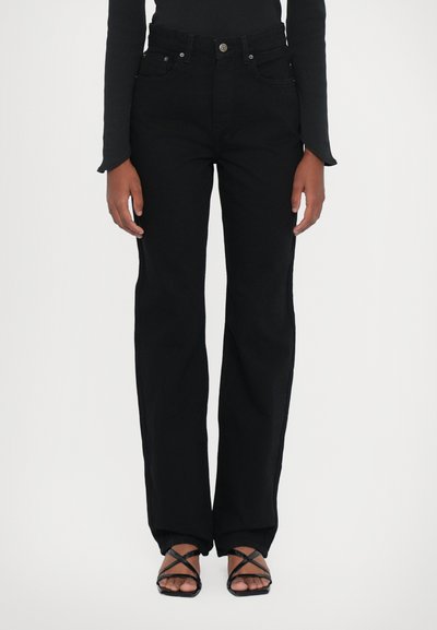 Jean noir taille haute avec une coupe droite. Comprend cinq poches, une fermeture à bouton et des détails de couture subtils. Assorti à des sandales noires à lanières.