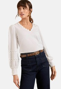 Top bianco con scollatura a V ondulata, maniche lunghe a palloncino e dettagli a occhio di pernice. Abbinato a jeans scuri a vita alta e una cintura marrone.