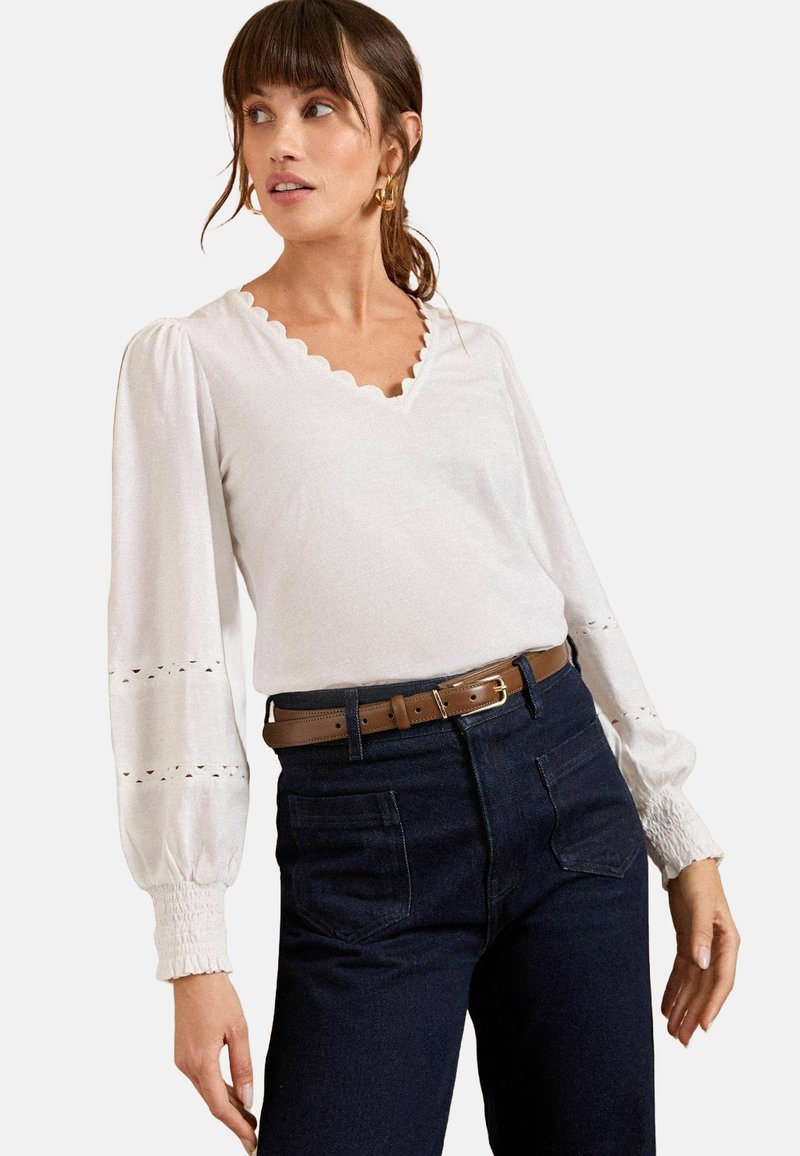 Top bianco con scollatura a V ondulata, maniche lunghe a palloncino e dettagli a occhio di pernice. Abbinato a jeans scuri a vita alta e una cintura marrone.