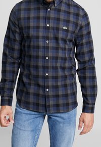Hombre con una camisa de cuadros en tonos azul, negro y gris, con un pequeño logo de cocodrilo verde, y jeans de un tono azul claro.