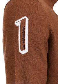 Pull en maille marron avec col roulé, arborant un "1" brodé en blanc et un détail en texte marine sur la manche gauche.