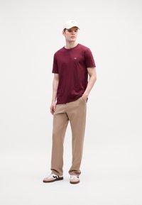 Jonge man in een bordeauxrood t-shirt, beige broek, witte sneakers en een lichtkleurige baseballcap, staande met één hand in zijn zak.