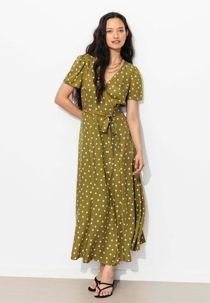 Femme portant une robe portefeuille vert olive à pois blancs, manches courtes, et sandales noires à lanières, debout contre un mur blanc.