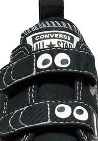 Zapatilla de lona negra con correas de velcro, costuras blancas y acentos circulares. Cuenta con la icónica marca Converse All Star en la lengüeta.