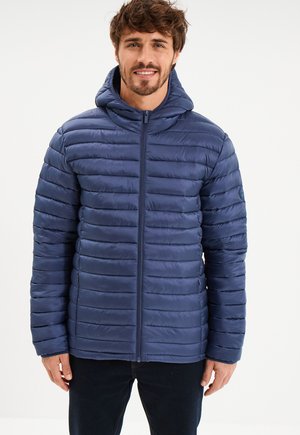 Mężczyzna ubrany w granatową, pikowaną kurtkę puchową z kapturem na zamek, stojący na tle plain białego tła, uśmiechający się.