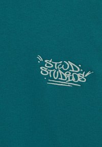 Logo ricamato "STUDIOS" in stile graffiti bianco su sfondo di tessuto teal.
