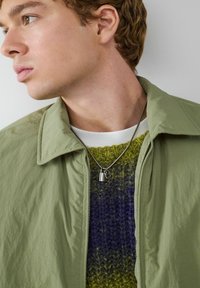 Jasnozielona kurtka z kołnierzykiem nałożona na dzianinowy sweter w odcieniach fioletu i zieleni, z srebrnym naszyjnikiem z małym wisiorkiem. Widoczne teksturowe powierzchnie.