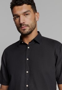 Zwarte button-up shirt van een gestructureerde stof, met korte mouwen en een kraag. Beschikt over lichtgekleurde knopen en een klein logo op de borst.