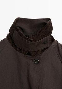 Braune Jacke mit einem kontrastierenden Kordkragen, Knopfverschluss und einer glatten Stofftextur, die ein modernes Designelement hervorhebt.