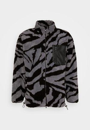 Chaqueta de forro polar con cremallera y cuello alto con rayas de cebra abstractas en negro y gris y un bolsillo en el pecho con cremallera negra en el lado izquierdo.