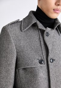 Grå rock med hundtandsmönster, spetsig krage, svarta knappar och texturerat tyg. Har axelklaff och en figurnära design.