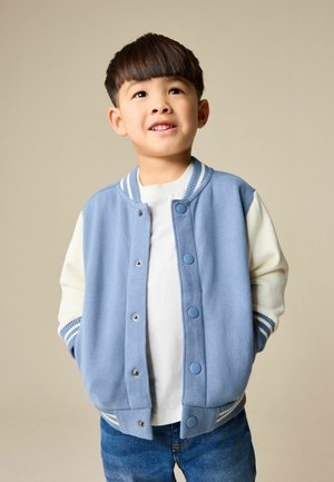 Blaues Fleece-Collegejackett mit weißen Ärmeln, gerippten Bündchen und Druckknöpfen. Trägt man über einem weißen Hemd, kombiniert mit blauen Jeans.