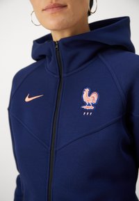 Tengerészkék Nike kapucnis pulóver, teljes cipzárral, kiemelkedő kakas emblémával és 'FFF' felirattal. Béléses kapucnival és sima anyagtextúrával rendelkezik.