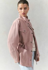 Veste surdimensionnée rose clair avec un col haut, des boutons-pression, des poches avant et un détail de taille froncée. Tissu doux et texturé.