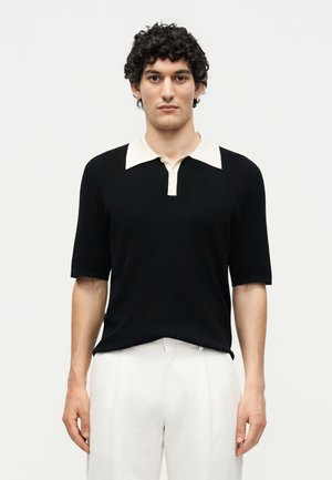 Άνδρας με κοντά σγουρά μαλλιά, που φοράει μαύρη κοντομάνικη μπλούζα πόλο με λευκό γιακά και λευκό παντελόνι, στέκεται μπροστά σε απλό φόντο.